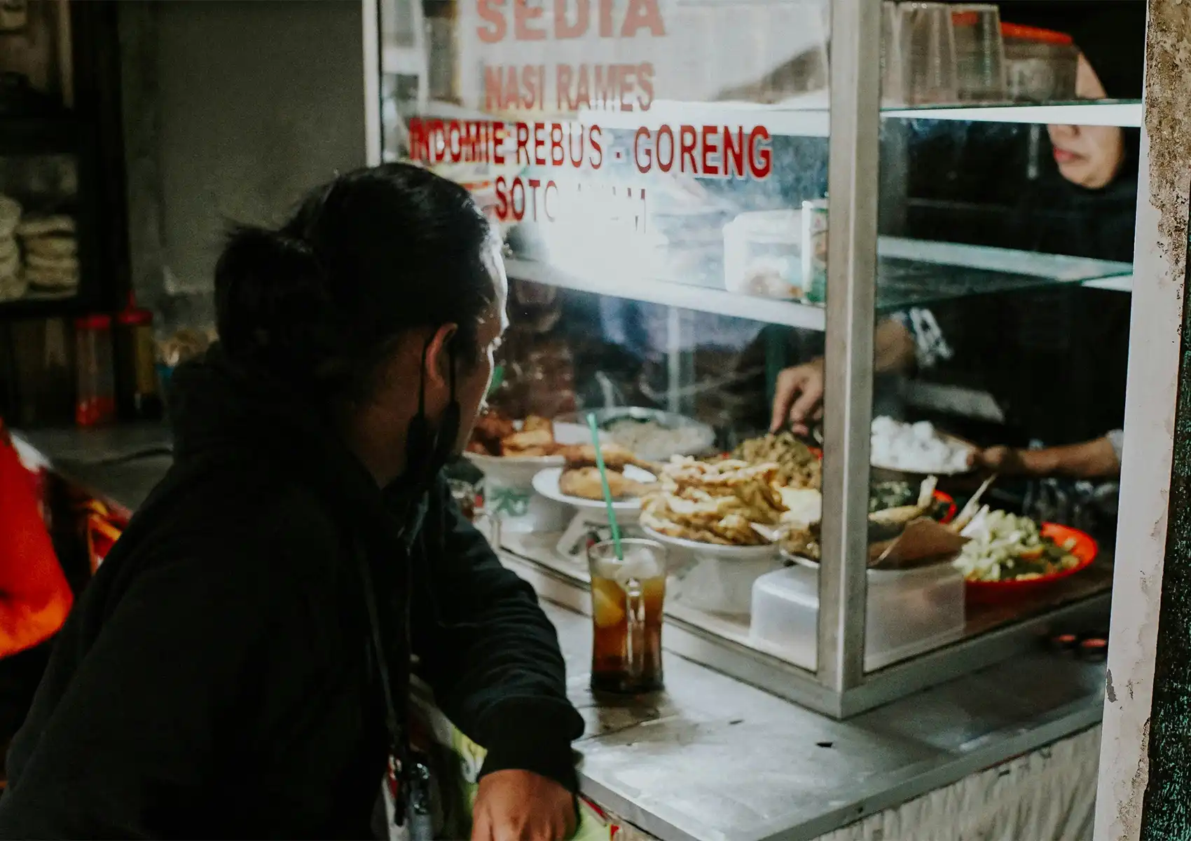 Indonesian street food stall serving nasi rames with fried dishes and iced tea to customers.