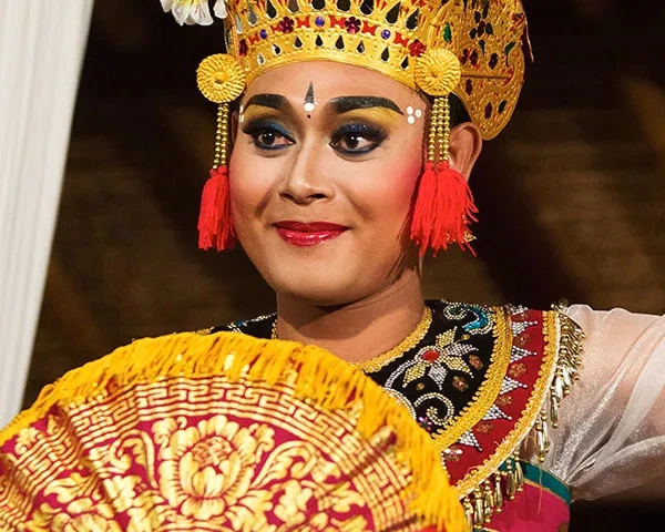 Balinese dancer in ornate golden headdress with fan, vibrant costume, and traditional makeup during cultural performance in Bali.