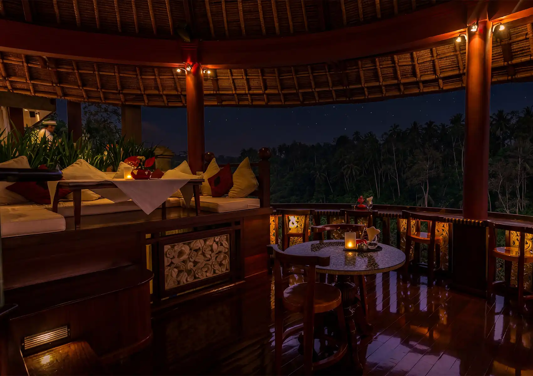 Cozy open‑air dining area in Bali with wooden furniture, candlelight, and jungle views under a starry night sky.