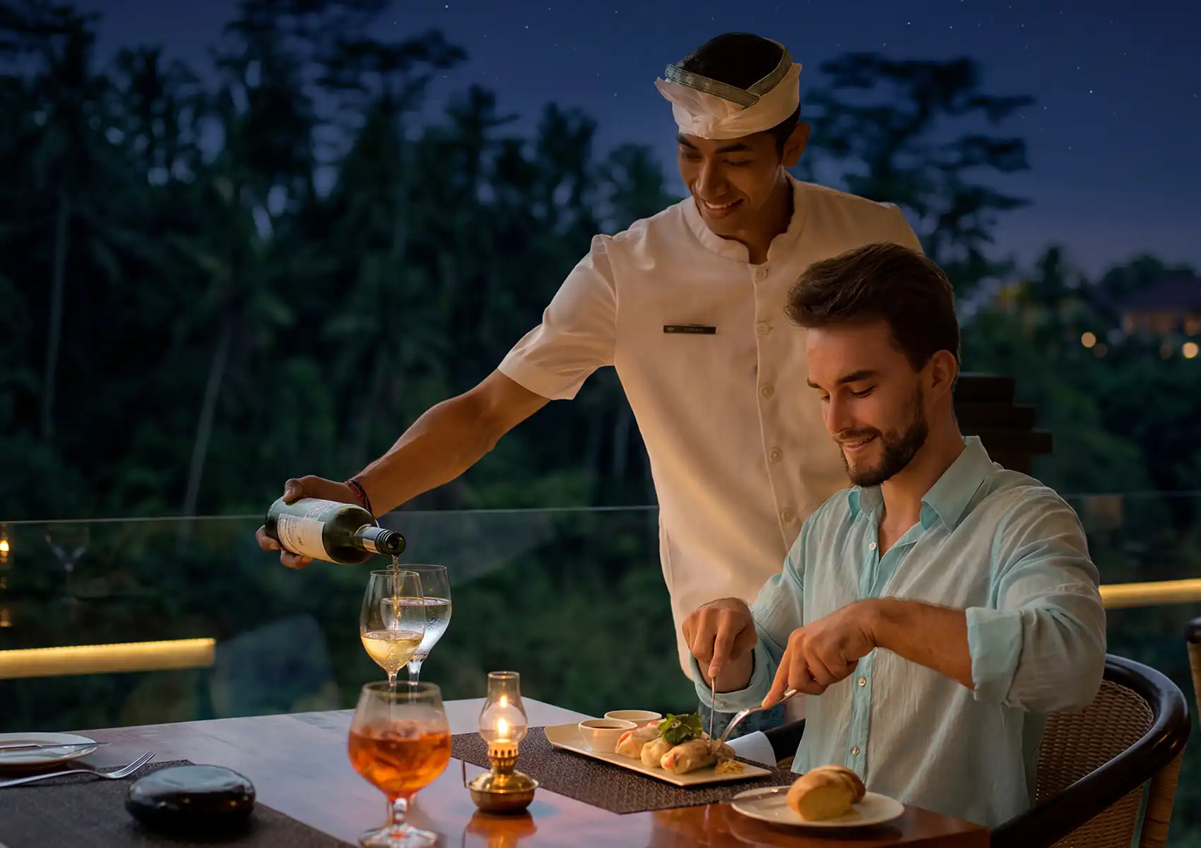 Outdoor restaurant in Ubud with server pouring wine under a starry sky, offering a refined dining experience.
