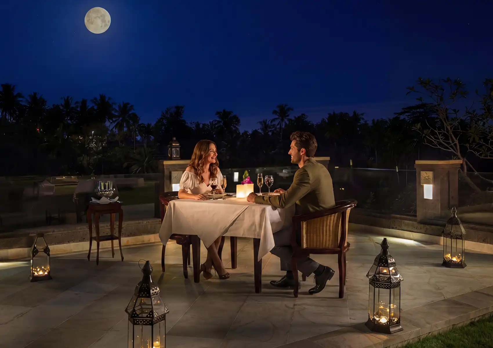 Romantic outdoor dinner under the full moon with candlelit table, wine glasses, and lanterns at an elegant Bali setting.