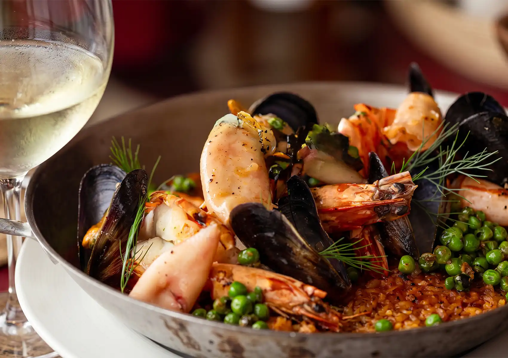 A close-up of a seafood paella with prawns, mussels, scallops, and green peas over saffron rice, served alongside a glass of white wine at a gourmet restaurant in Ubud, Bali.