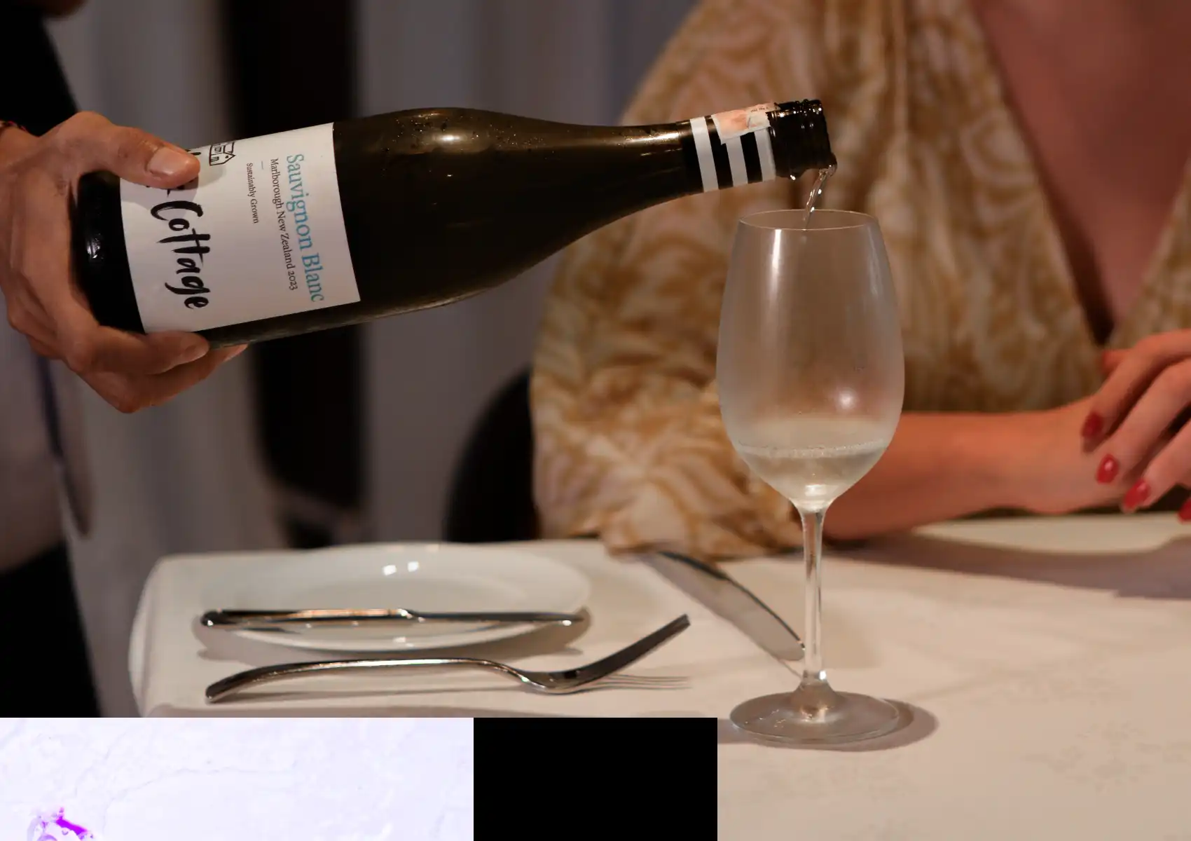 A server pours a glass of New Zealand Sauvignon Blanc white wine for a diner at a restaurant in Ubud, Bali, with elegant table settings of white plates and polished silverware on a white tablecloth.