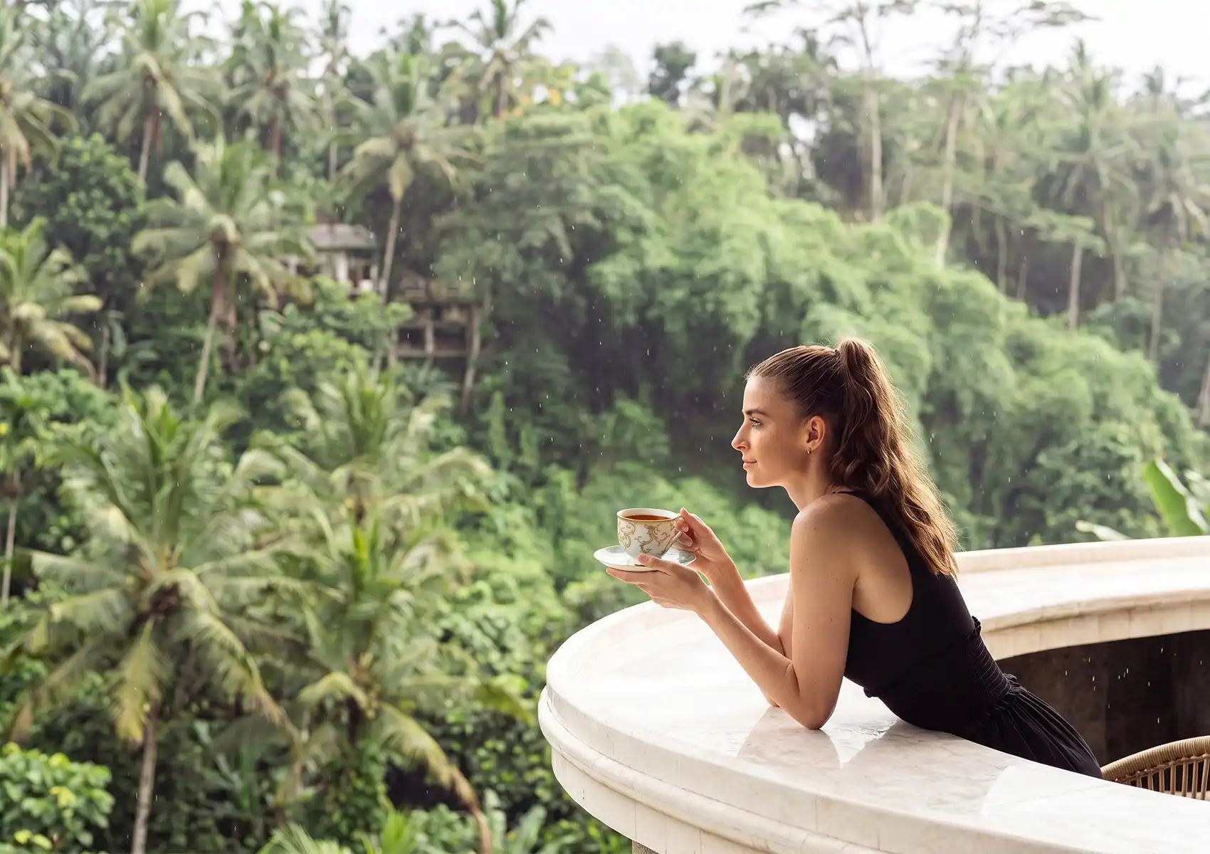 A woman in a black top leans on a curved marble balcony railing while holding a delicate teacup, gazing out over a lush palm-filled jungle valley at a restaurant in Ubud, Bali.