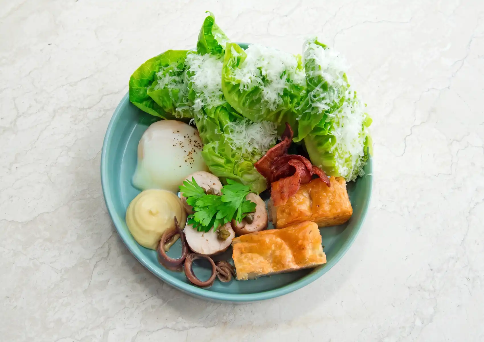 An overhead view of a deconstructed Caesar salad served at a restaurant in Ubud, on a teal plate featuring butter lettuce topped with grated parmesan, a soft poached egg, crispy bacon, golden croutons, rolled chicken, anchovies, capers, and a dollop of mayonnaise.