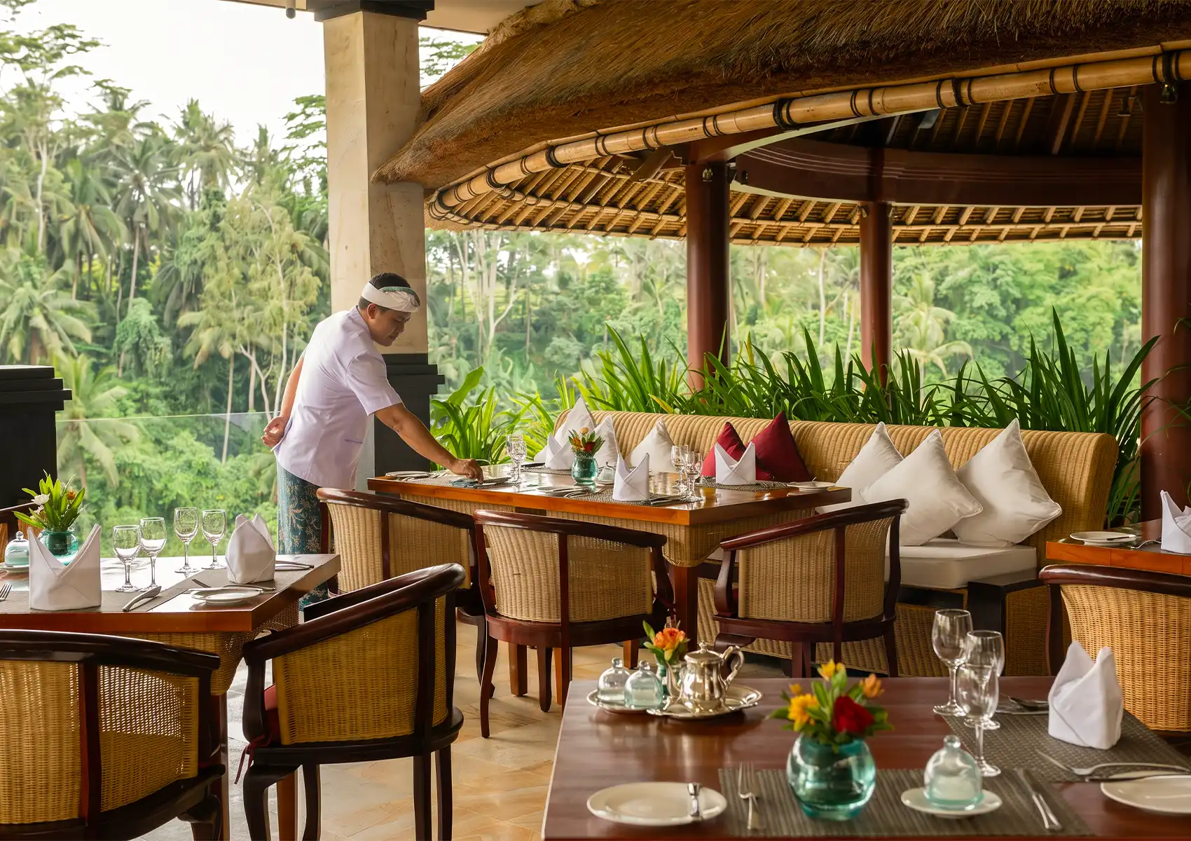 A waiter in a white uniform and headband sets a table at a restaurant in Ubud with a thatched roof, wicker furniture, and white linens overlooking a lush green jungle in Bali.