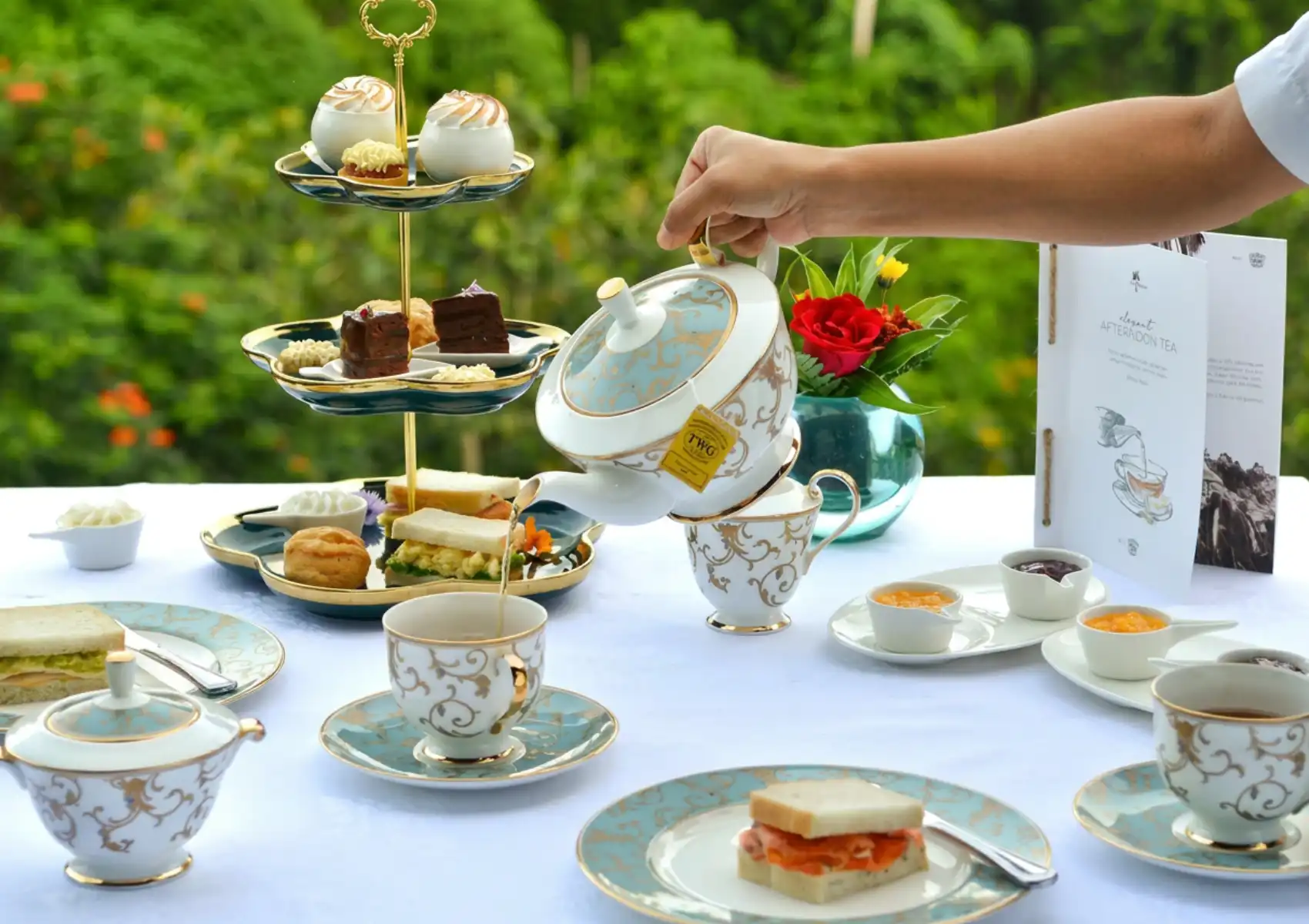A hand pours tea from an ornate gold and white TWG teapot into a matching cup at a restaurant in Ubud, with a tiered stand of finger sandwiches, scones, chocolate cakes, and meringues set on a white tablecloth against lush greenery.