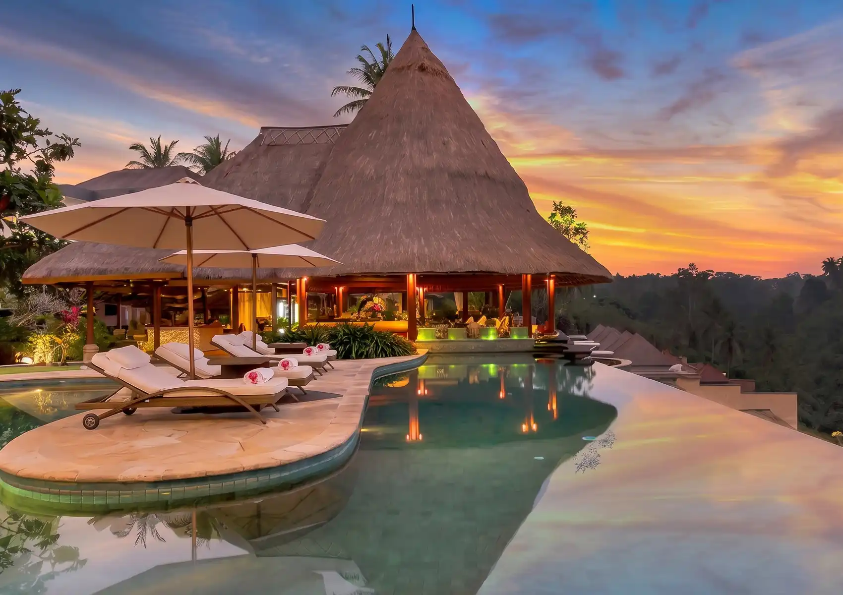 A luxury infinity pool with sun loungers and umbrellas beside a thatched-roof pavilion at a restaurant in Ubud, glowing warmly against a vibrant pink and orange sunset over the jungle.