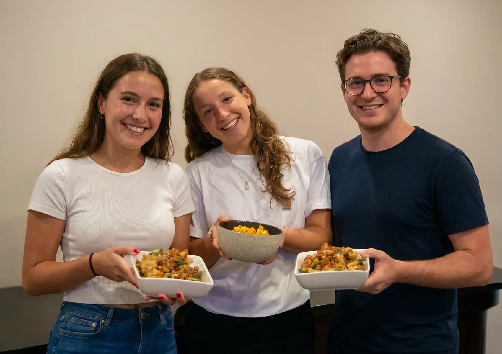 Three smiling young adults holding bowls of freshly prepared food, showcasing the delicious results of their hands-on cooking class in ubud.