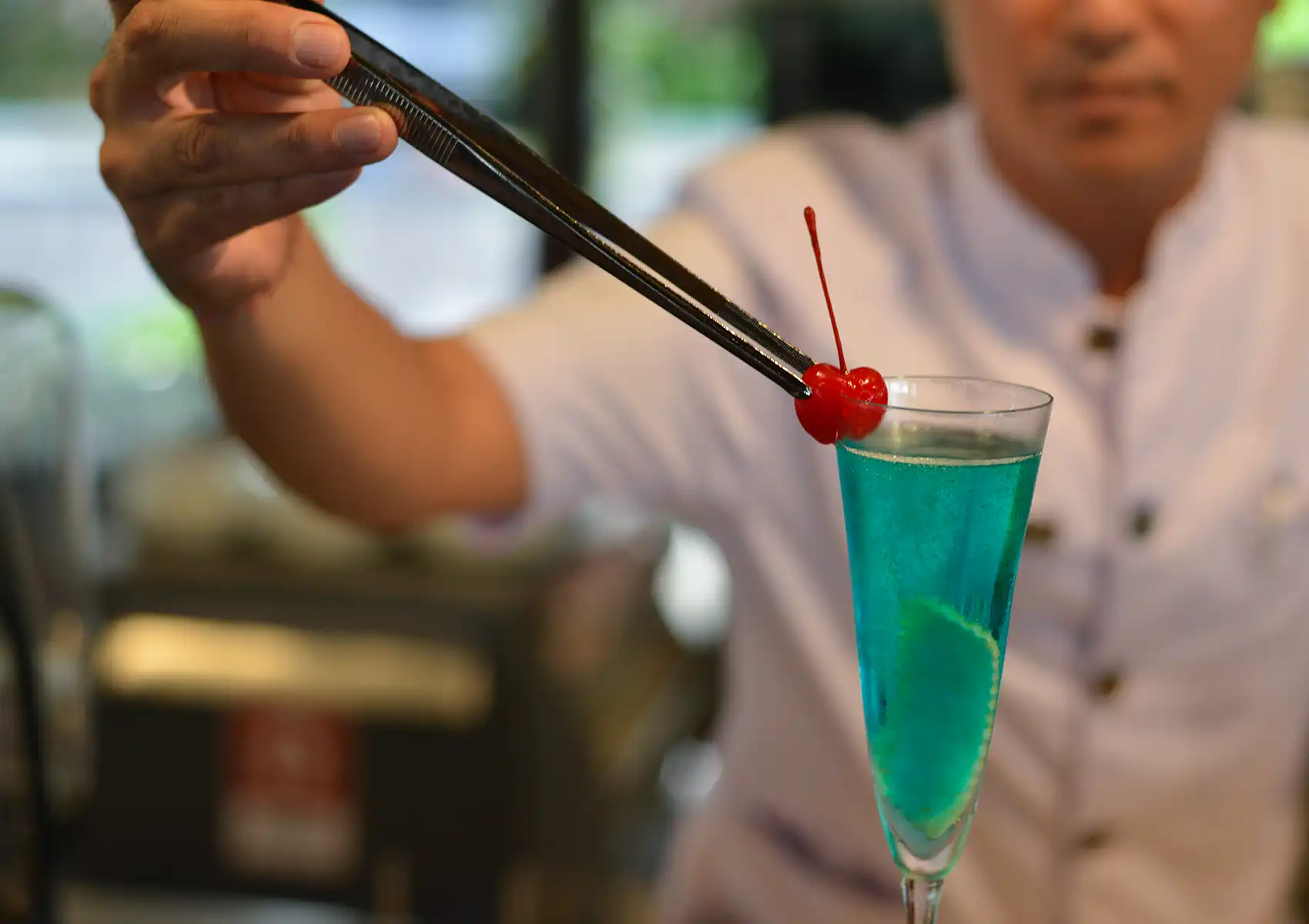 A close-up of a bartender carefully using long metal tweezers to garnish a vibrant blue drink in a tall glass with a bright red cherry, showcasing the expert mixology behind tropical cocktails ubud.