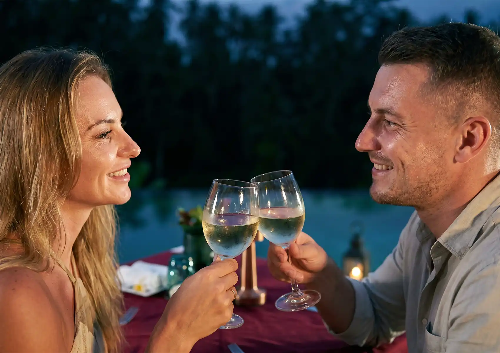 A close-up of a happy couple looking into each other's eyes and toasting with glasses of white wine across a table set with a red cloth, celebrating a romantic evening with a seven course dinner ubud.