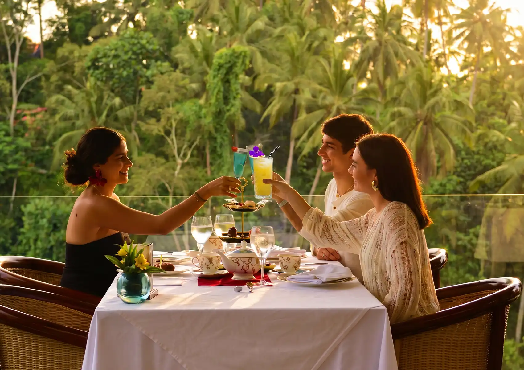 Three friends smile and toast with colorful tropical cocktails ubud at a beautifully set dining table, enjoying their drinks against the stunning backdrop of a lush, sunlit green jungle valley.