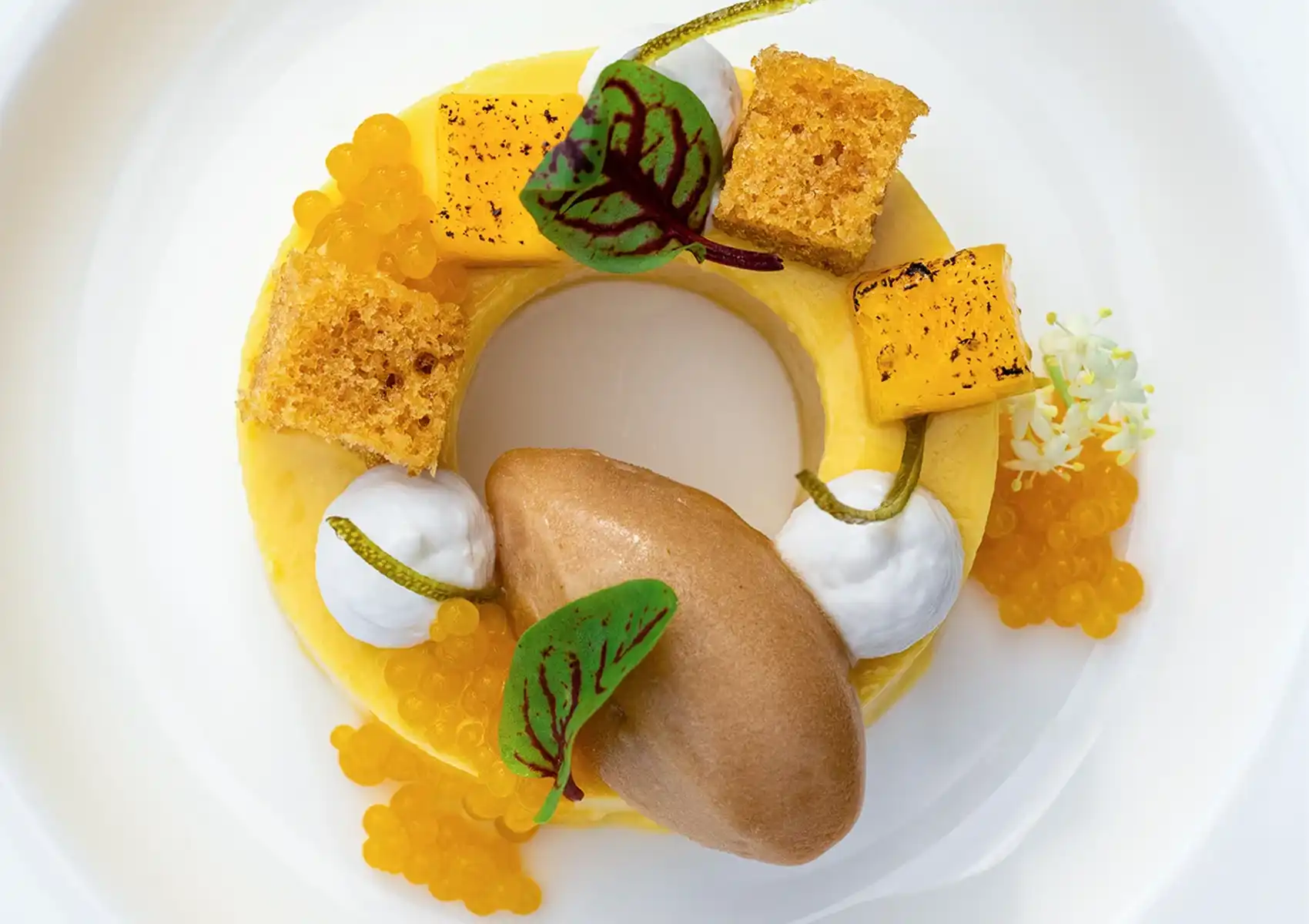 A beautifully plated gourmet dish featuring a vibrant yellow base, delicate pieces of sponge cake, a smooth quenelle, dollops of white cream, and bright orange roe on a white plate, showcasing the culinary artistry of a seven course dinner ubud.