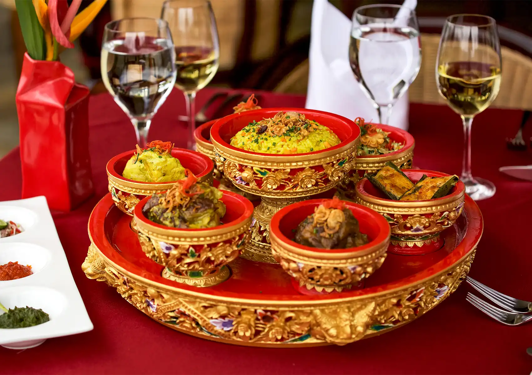 An elegant spread of traditional Balinese cuisine featuring yellow rice and various side dishes in ornate red and gold bowls, served with glasses of white wine at a fine dining restaurant in Ubud.