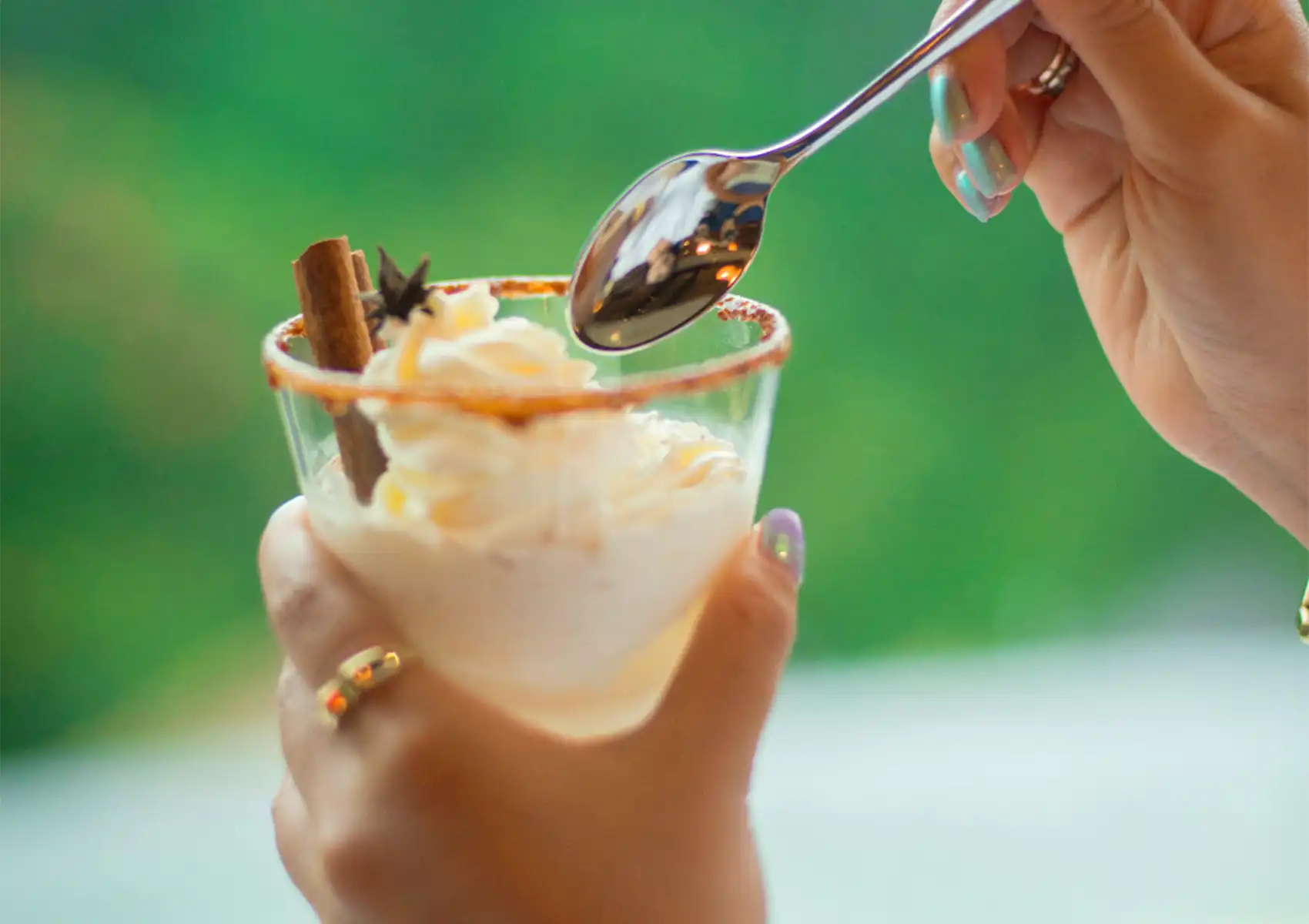 A hand holding a creamy glass dessert topped with whipped cream and a cinnamon stick against a blurred, lush green background at a restaurant in Ubud.