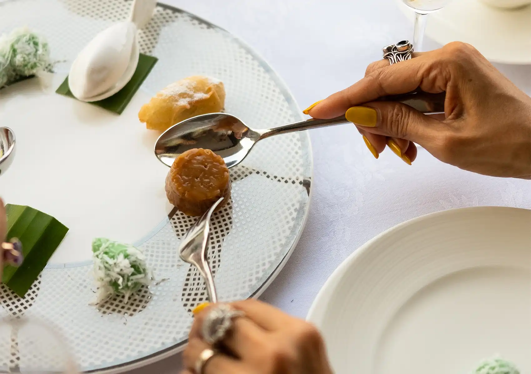 A close-up of a person using a spoon and fork to taste a selection of traditional bite-sized sweet treats served on small banana leaves at a restaurant in Ubud.