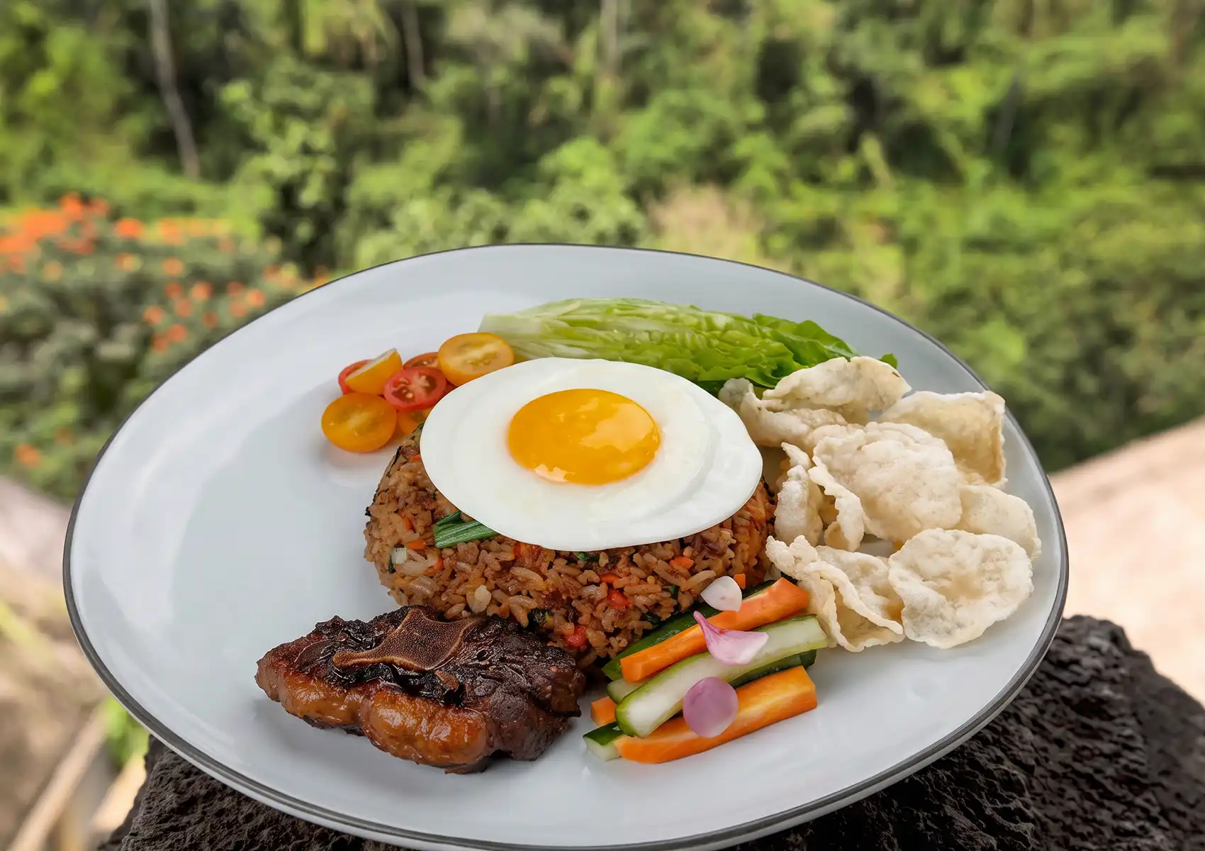 A beautifully plated dish of Indonesian Nasi Goreng topped with a sunny-side-up egg, accompanied by grilled meat and crackers, set against a lush green tropical backdrop at a restaurant in Ubud.