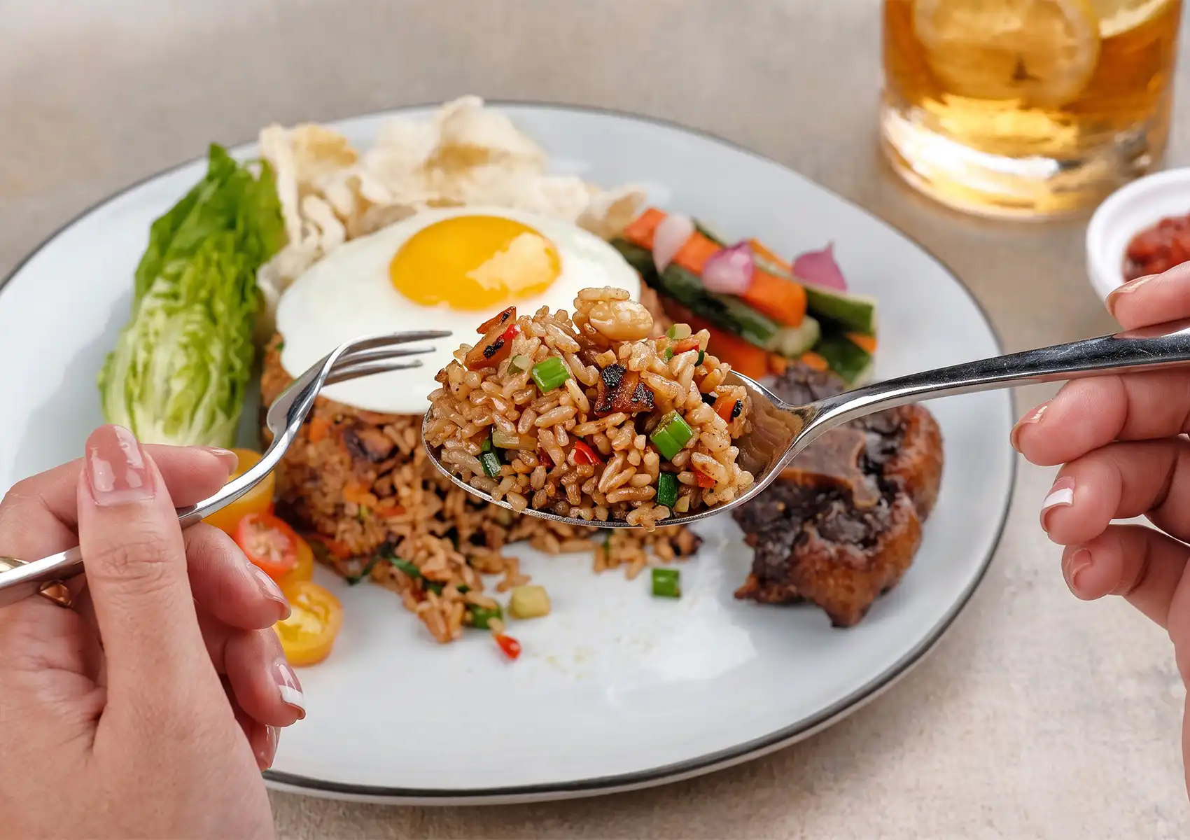A close-up of a spoonful of savory Indonesian fried rice being lifted from a plate featuring a fried egg, grilled meat, and crackers at a restaurant in Ubud.