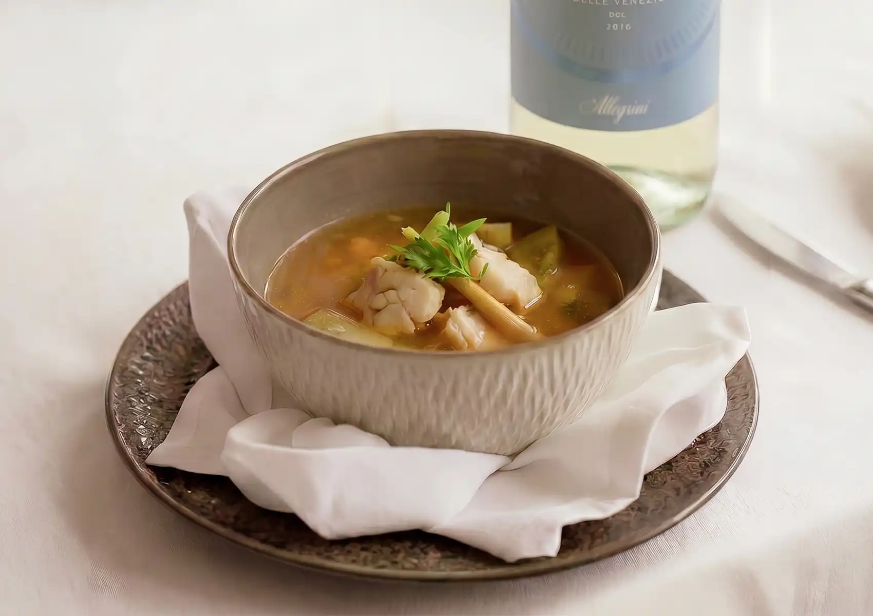 An elegant presentation of Kuah Be Pasih, a traditional Balinese clear fish soup garnished with lemongrass, served alongside a bottle of white wine at a fine dining restaurant in Ubud.