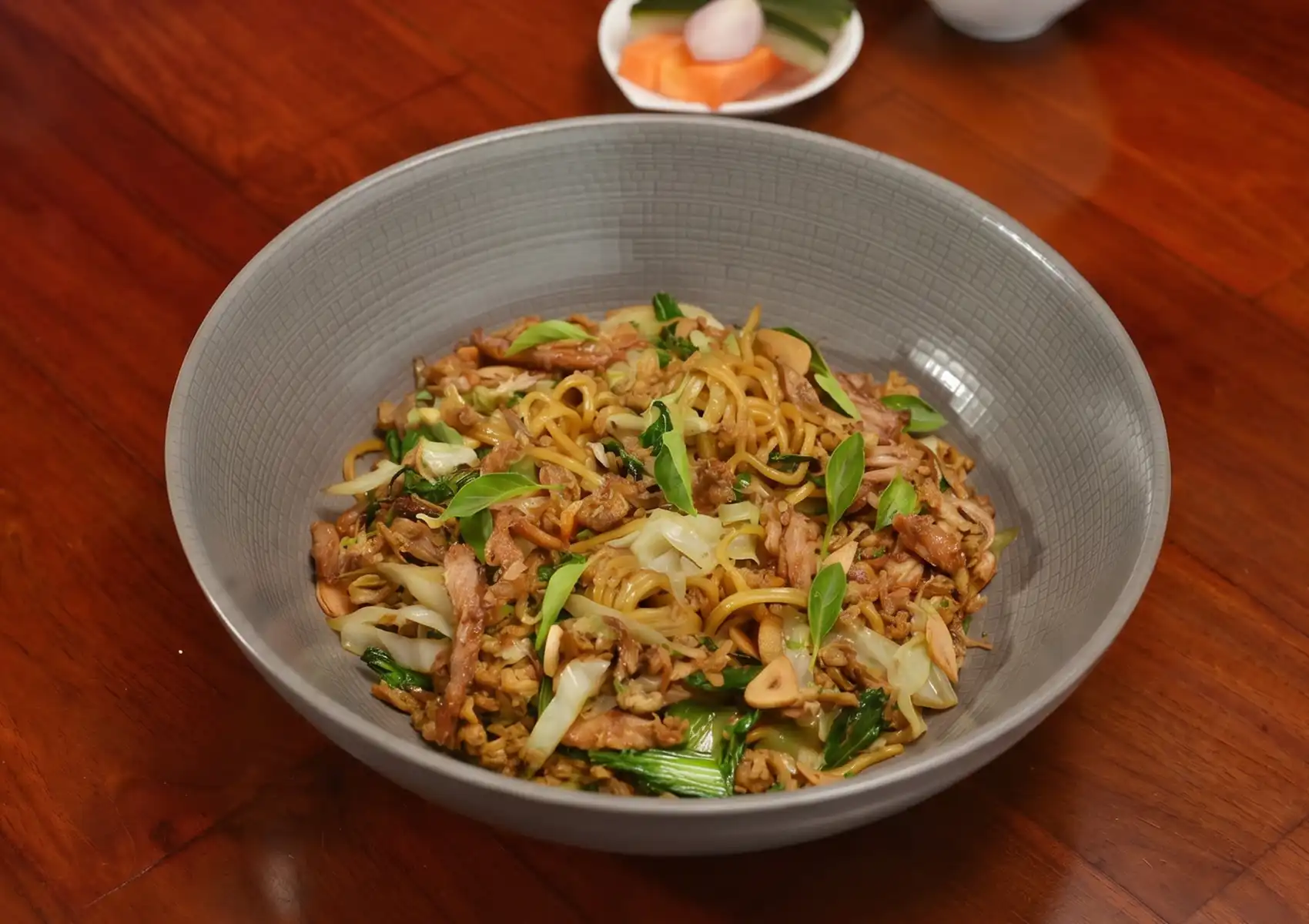A close-up overhead view of Mie Goreng Cakalang, a savory dish of Indonesian fried noodles tossed with vegetables and herbs, served in a grey bowl at a restaurant in Ubud.