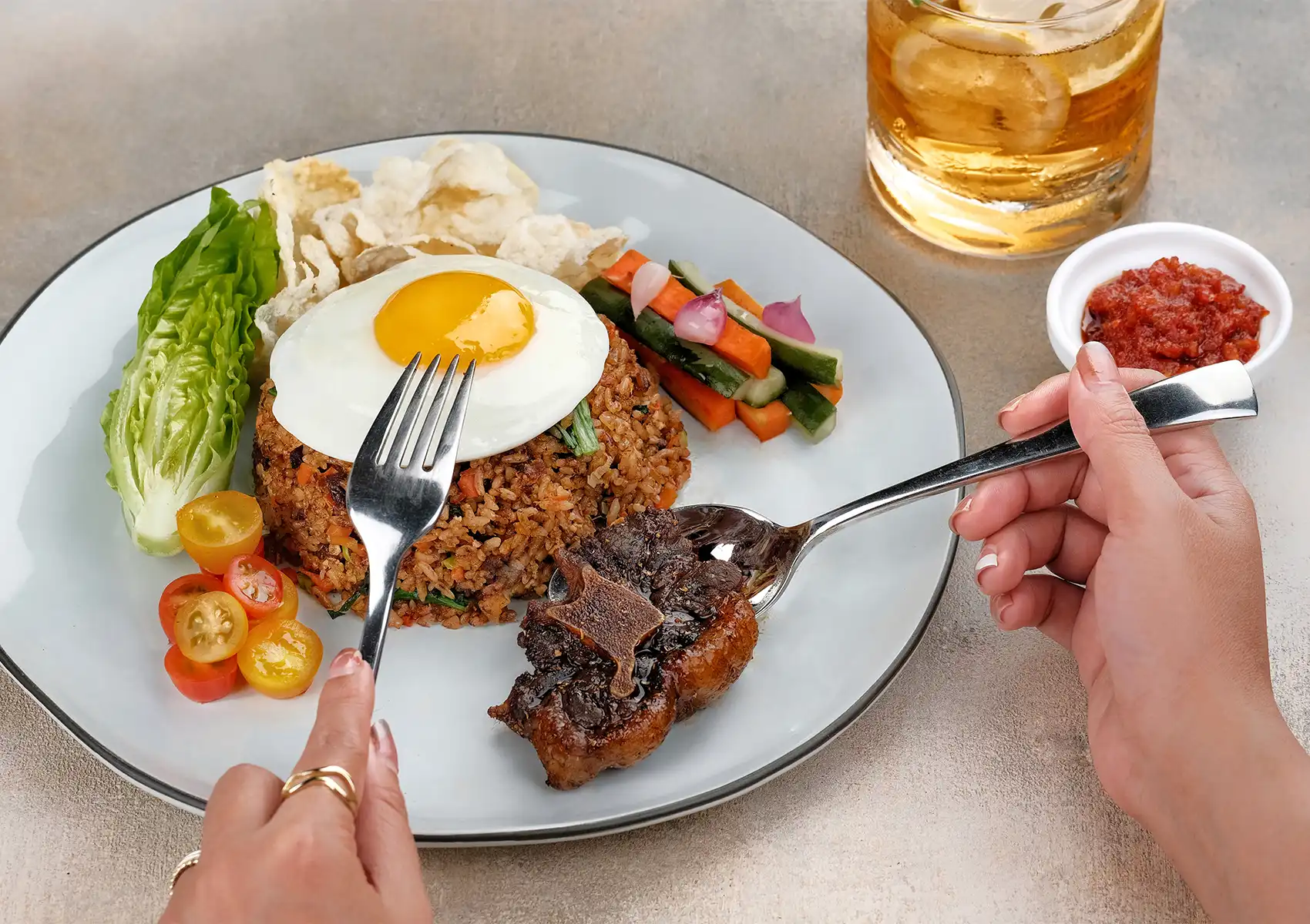 A close-up of a diner using a spoon to lift a piece of glazed meat from a traditional Nasi Goreng dish served with a fried egg and fresh vegetables at a restaurant in Ubud.