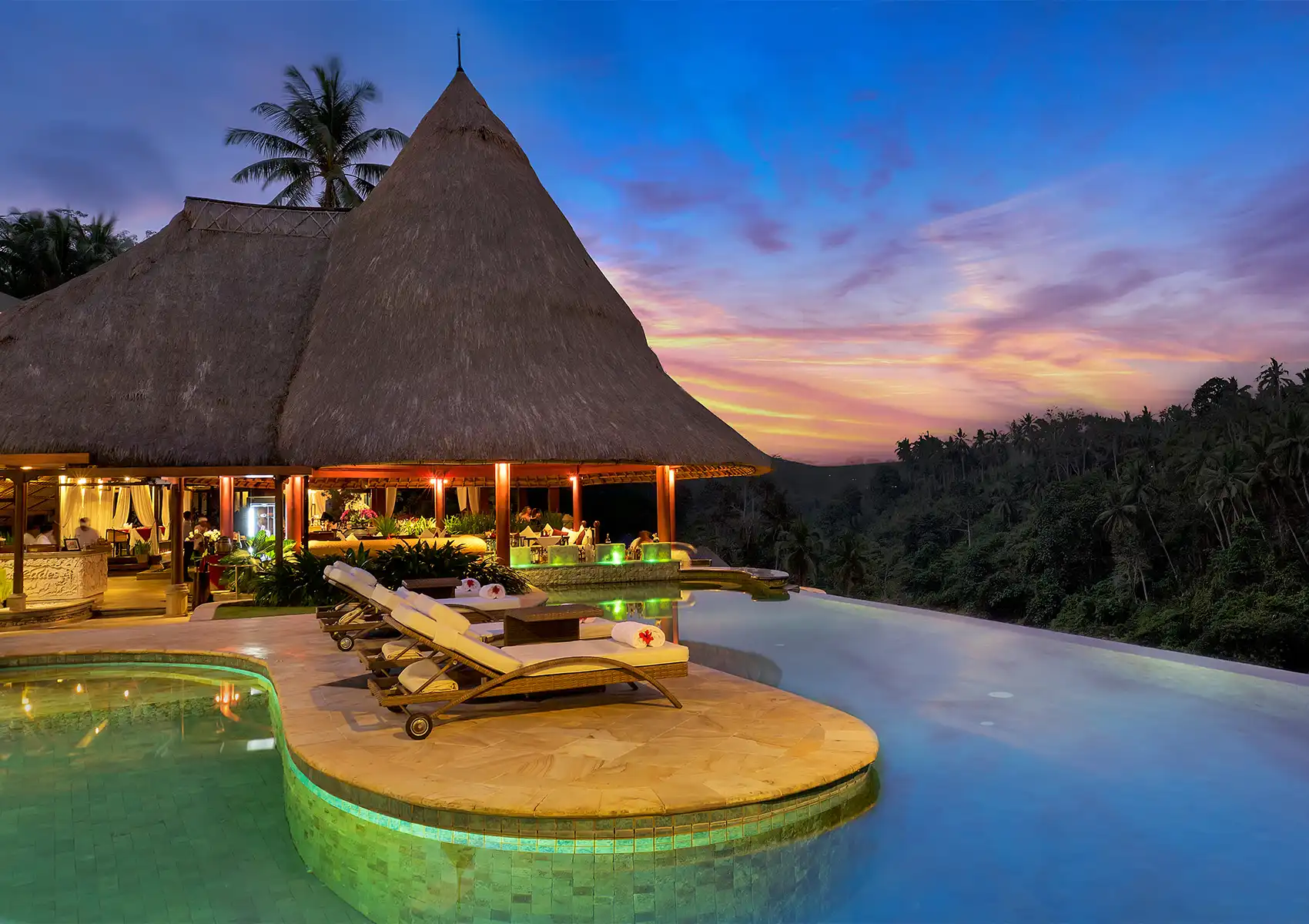 A stunning thatched-roof open-air restaurant in Ubud glowing warmly at twilight, situated right next to a luxurious infinity pool overlooking a lush tropical jungle valley.