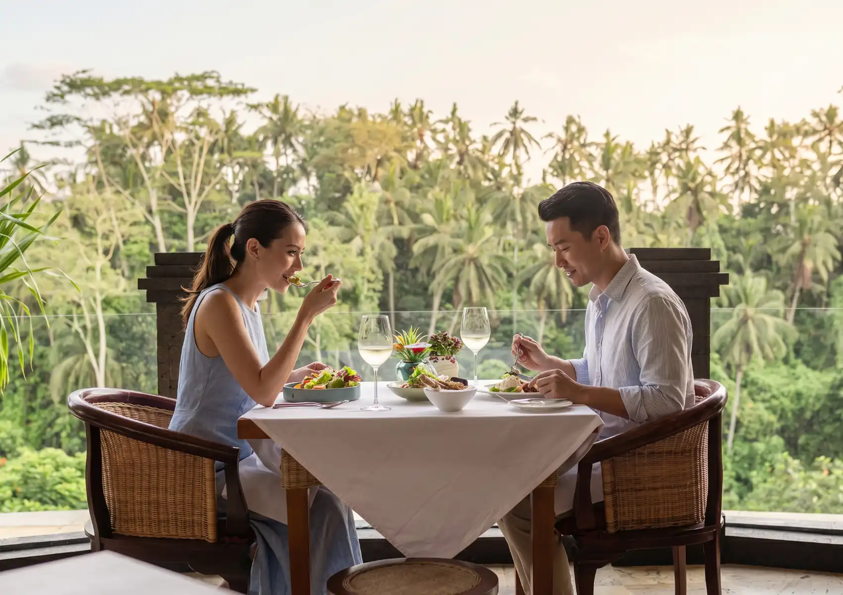 A couple eating lunch together at a beautifully set table on a balcony at a restaurant romantic Ubud, featuring a scenic backdrop of palm trees and tropical forest