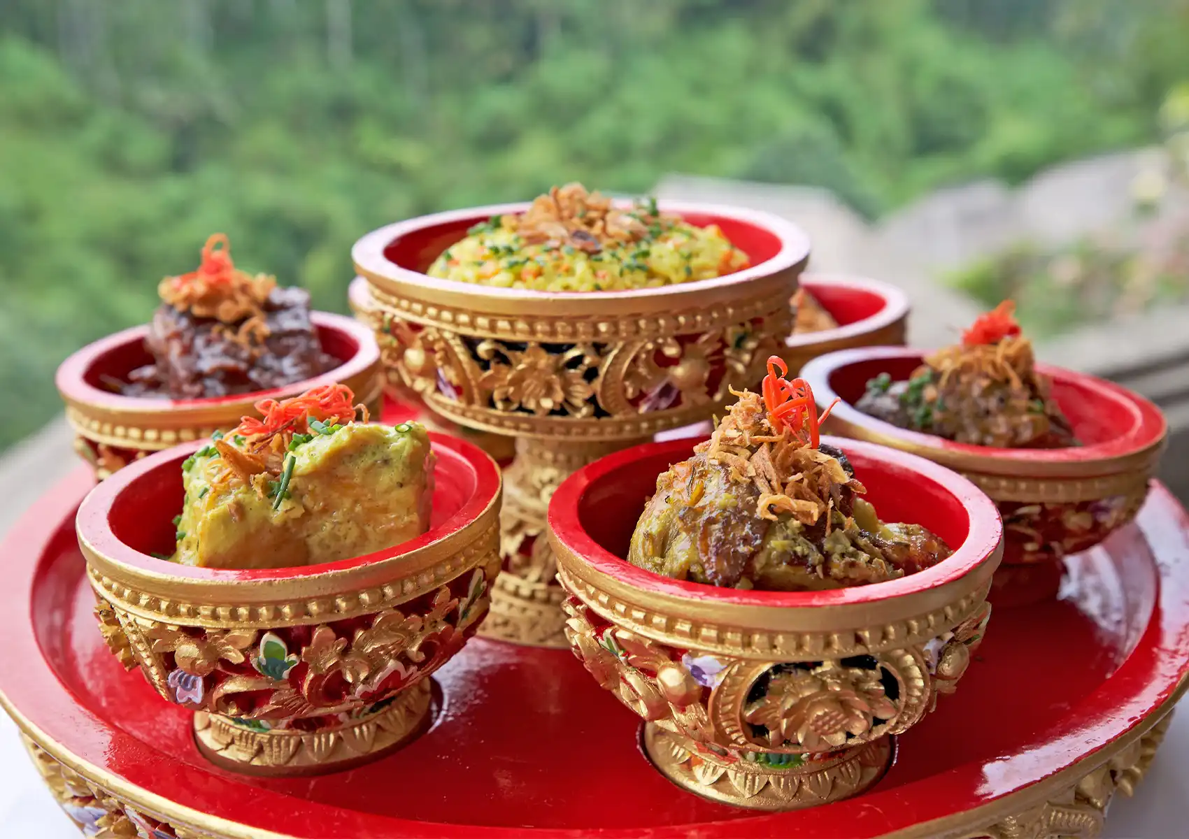 A traditional, ornate red and gold tiered tray presenting an assortment of Indonesian sharing dishes against a lush green valley backdrop at a restaurant in ubud.