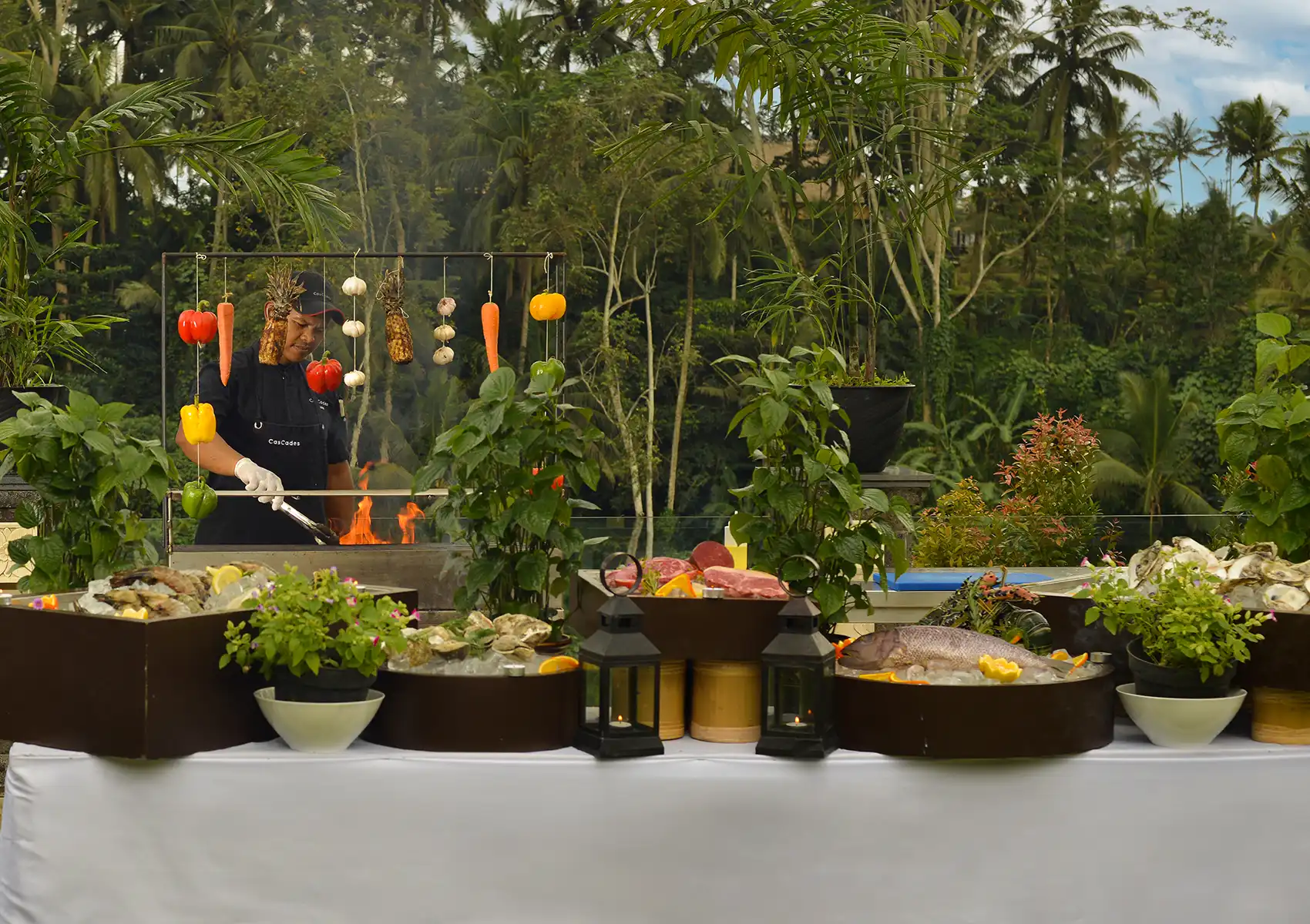 A chef tends to an open fire grill behind a lavish buffet of fresh seafood, steaks, and vegetables displayed on ice outdoors at a Restaurant in ubud.
