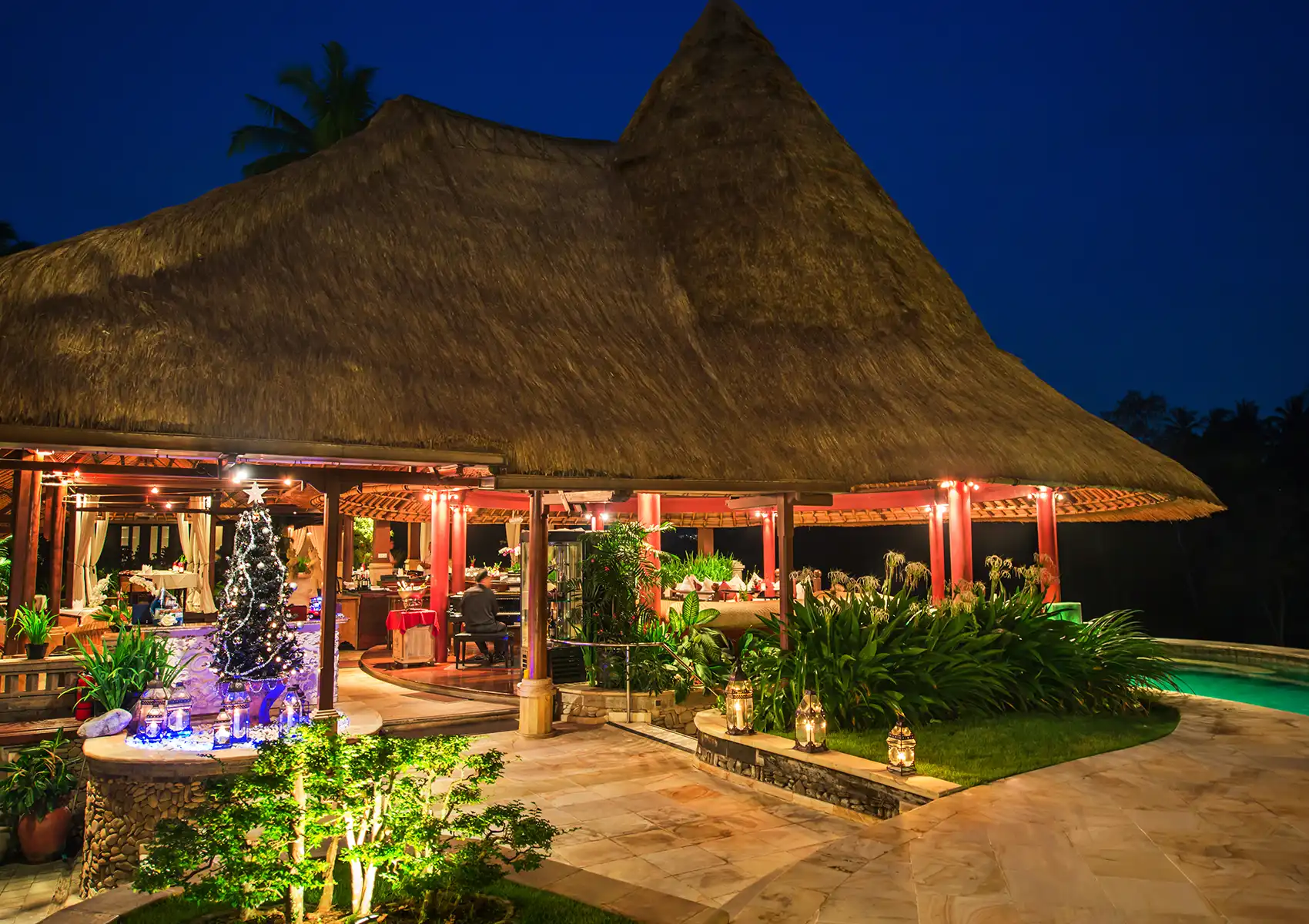 A warmly illuminated, traditional thatched-roof Restaurant in ubud at night, featuring a decorated Christmas tree and glowing lanterns by the entrance for a festive holiday celebration.