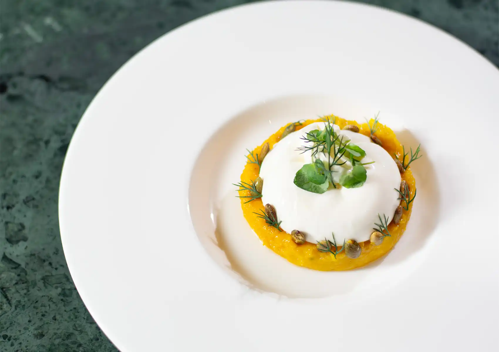 An elegant appetizer featuring a smooth sphere of creamy burrata cheese placed atop a vibrant orange puree, garnished with fresh dill and toasted pumpkin seeds on a pristine white plate at a Restaurant in ubud.