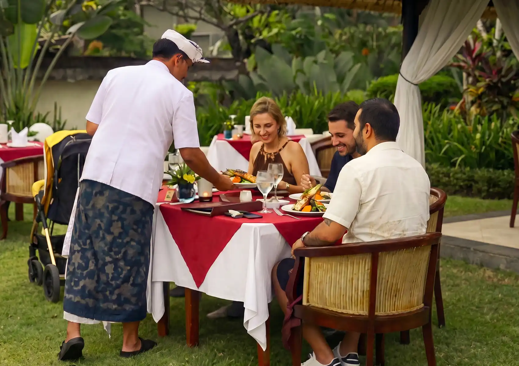 A waiter serves a plated grilled dinner to a smiling group of friends seated at an elegantly set table on a grassy lawn at a Restaurant in ubud.