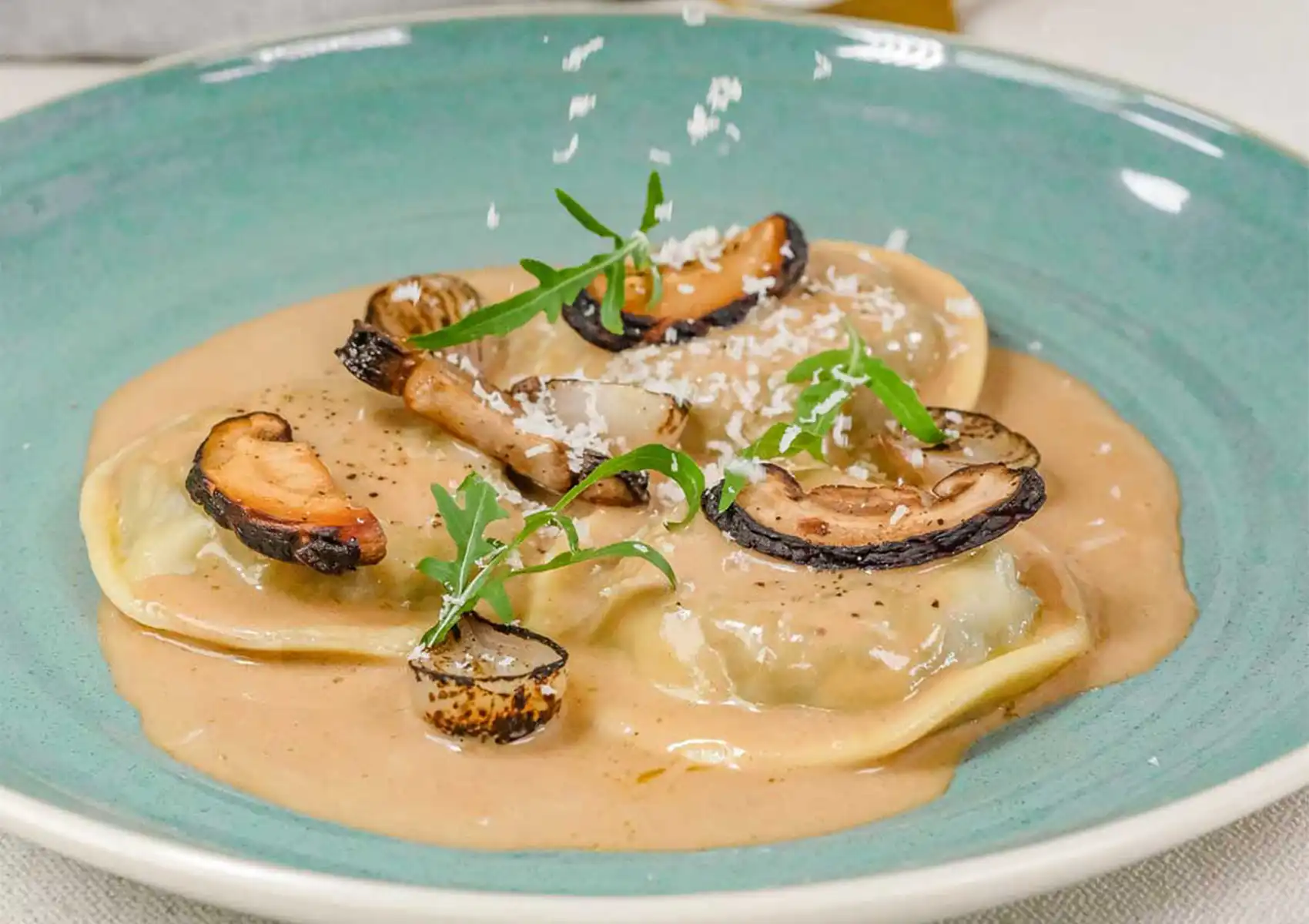A chef sprinkles fresh parmesan cheese over a plate of delicate lobster ravioli, served in a creamy sauce and topped with sliced mushrooms and arugula at a Restaurant in ubud.