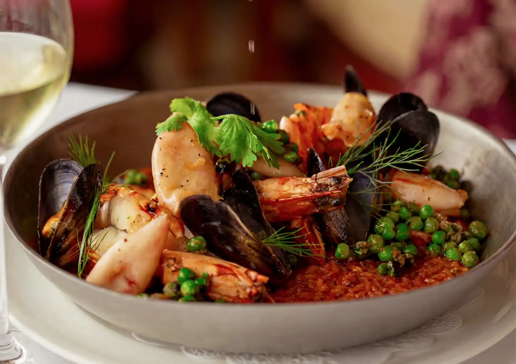 A beautifully plated bowl of savory seafood paella loaded with fresh mussels, grilled prawns, calamari, and green peas, accompanied by a glass of white wine at a Restaurant in ubud.