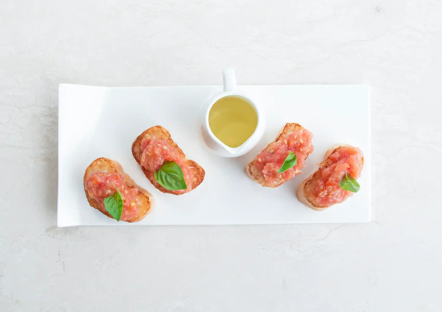 A top-down view of four pieces of fresh tomato bruschetta garnished with vibrant green basil leaves, served alongside a small pitcher of olive oil on a sleek white plate at a Restaurant in ubud.